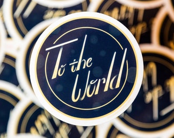 May include: Close-up of a round sticker with a gold circle and the text "To the World" in a gold script font on a dark blue background.