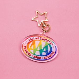 May include: A white oval keychain with a rainbow-colored "4A" logo and the text "Call Me Triple "A" Anxious + ADHD + Autistic". The keychain has a gold star-shaped charm.