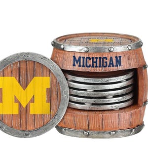May include: A wooden barrel-shaped coaster holder with the word "MICHIGAN" in navy blue. The holder contains several silver coasters. A separate coaster with a yellow "M" logo is also visible.