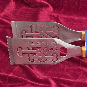 May include: Stainless steel grilling tongs with wooden handles. The tongs feature the New York Mets logo and text. A blue band with the words "Grill-A-Tongs" is wrapped around the handles.