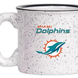May include: White ceramic mug with a speckled pattern and a black rim. The mug features the Miami Dolphins logo, including a leaping dolphin in teal and orange, and the team name in teal.