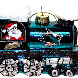May include: A teal and black train ornament featuring the San Jose Sharks logo and a Santa Claus graphic. The ornament has a glittery finish and a gold-colored metal loop for hanging. The wheels are silver and black.