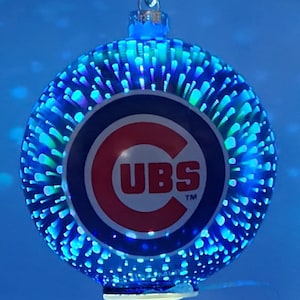 Pode incluir: Uma bola de Natal redonda e iluminada com o logotipo do Chicago Cubs. O enfeite apresenta um logotipo azul e vermelho com a palavra "CUBS" em branco. O enfeite tem um padrão de luz azul e verde.