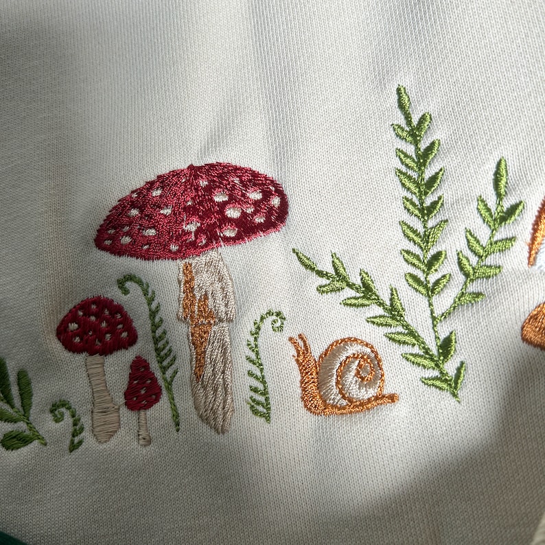 Op de afbeelding: Borduurwerk met paddenstoelen en een slak op een witte stoffen achtergrond. De paddenstoelen zijn rood met witte stippen en hebben bruine stelen. De slak is bruin en heeft een schelp. Het ontwerp is omgeven door groene bladeren.