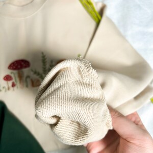 Op de afbeelding: Een close-up van een cr&egrave;mekleurig sweatshirt met geborduurde paddenstoelen en bladeren. De stof is zacht en heeft een textuur.