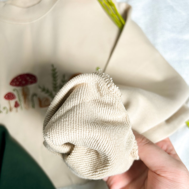 Op de afbeelding: Een close-up van een cr&egrave;mekleurig sweatshirt met geborduurde paddenstoelen en bladeren. De stof is zacht en heeft een textuur.