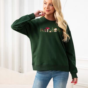 Op de afbeelding: Een donkergroen sweatshirt met een geborduurd ontwerp van paddenstoelen en bladeren. Het sweatshirt heeft een ontspannen pasvorm en een ronde hals.