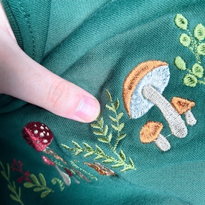 Op de afbeelding: Groen stof met geborduurde paddenstoelen in tinten van oranje, bruin en wit. De paddenstoelen worden omringd door groene bladeren.