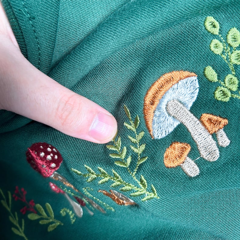 Op de afbeelding: Groen stof met geborduurde paddenstoelen in tinten van oranje, bruin en wit. De paddenstoelen worden omringd door groene bladeren.