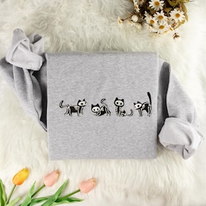 May include: Grey sweatshirt with a line of five black skeleton cats printed on the front. The cats are in various poses, including sitting, standing, and walking.