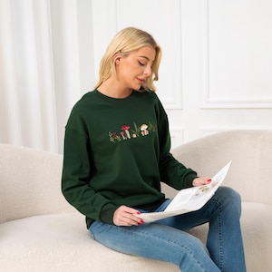 Op de afbeelding: Een donkergroen sweatshirt met een geborduurd ontwerp van paddenstoelen en bladeren. Het model draagt ​​een blauwe jeans en zit op een witte bank.