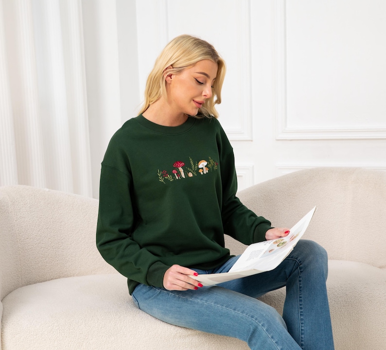 Op de afbeelding: Een donkergroen sweatshirt met een geborduurd ontwerp van paddenstoelen en bladeren. Het model draagt ​​een blauwe jeans en zit op een witte bank.
