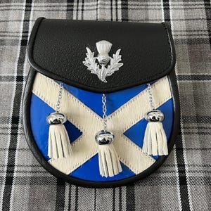Könnte beinhalten: Eine schwarze Leder-Sporran mit blau-weißem Design, silbernem Distelabzeichen und drei weißen Quasten. Die Sporran hat einen Klappenverschluss und eine Kette. Das Design ähnelt der schottischen Flagge.