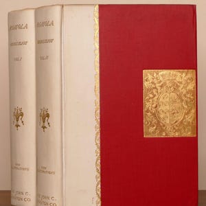 May include: Two volumes of Romola by George Eliot, published by The John C. Winston Co. The books feature white spines with gold lettering and a red cover with a gold embossed crest. The spines are labeled Vol. I and Vol. II with gold fleur-de-lis.