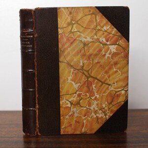 Könnte beinhalten: Ein antikes, braunes, ledergebundenes Buch mit goldener Beschriftung auf dem Rücken, die "Selections from Greek Anthology" lautet. Der Einband ist marmoriert in Orange, Gelb und Weiß.