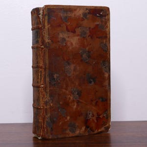 Puede incluir: Un libro antiguo con una cubierta de cuero marrón desgastada. La cubierta tiene un aspecto moteado con manchas más oscuras y desgaste visible. El lomo muestra bandas en relieve y signos de edad. El libro está de pie sobre una superficie de madera.