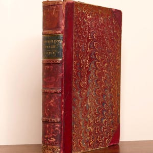 May include: A vintage hardback book titled A Naturalist's Voyage by Darwin, featuring a dark red leather spine with gold lettering and a marbled red, blue, and gold patterned cover with leather corners.