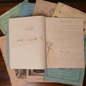 May include: A collection of vintage paper notebooks with soft covers in shades of green, blue, and beige. One open notebook displays handwritten French text titled De la vigne and a hand-drawn illustration of a person wearing a cap.