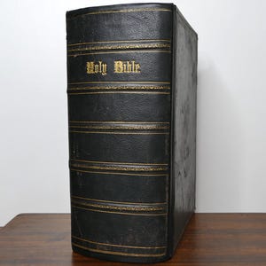 May include: A large, antique black leather-bound book with gold lettering that reads "Holy Bible." The book has gold accents and is standing upright on a wooden surface.