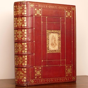 May include: A red leather-bound book with gold-embossed floral patterns on the spine and cover. The front cover features a central rectangular inlay depicting praying hands, with the text HOLY HOLY HOLY at the top and LORD GOD OF HOSTS at the bottom.
