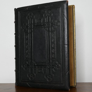 May include: A large, antique book with a black leather cover featuring an embossed decorative design. The book's spine is visible, and the pages have a gold-colored edge. The book is resting on a wooden surface.