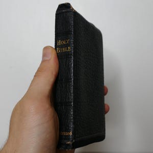 May include: A black, leather-bound Holy Bible with gold lettering. The book is held in a hand, showcasing the spine with the title "Holy Bible" and the word "Illustrated". The book's edges show signs of wear.