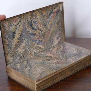 May include: An antique book with marbled paper covers and endpapers. The open book displays a decorative pattern in shades of brown, blue, and red. The book's pages are aged, and the binding is visible.