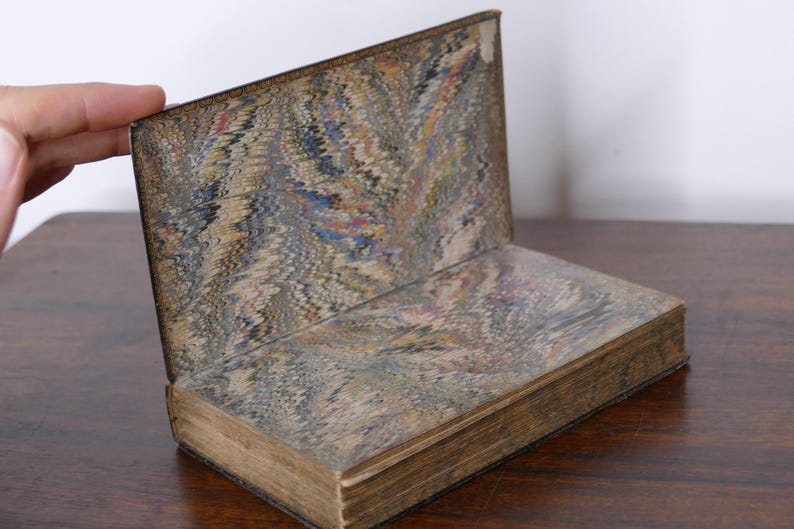 May include: An antique book with marbled paper covers and endpapers. The open book displays a decorative pattern in shades of brown, blue, and red. The book's pages are aged, and the binding is visible.
