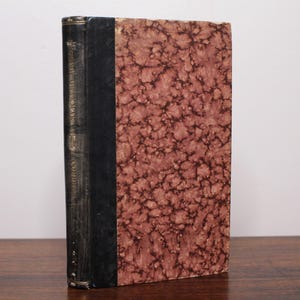 Könnte beinhalten: Ein Vintage-Hardcover-Buch mit schwarzem Rücken und marmoriertem Papierumschlag in Braun- und Rottönen.