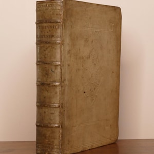 May include: A thick, antique book with a weathered, light brown leather cover. The spine features raised bands and faint, partially legible text. The front cover displays a subtle, embossed floral design in the center, showing signs of age.