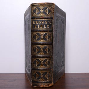 Puede incluir: Un libro antiguo con una cubierta de cuero oscuro. El lomo presenta el título "BROWN'S BIBLE" en letras doradas, con detalles dorados decorativos. El libro se exhibe sobre una superficie de madera.