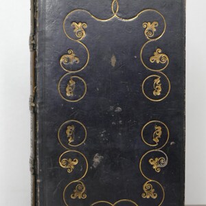 May include: Antique book with a dark blue leather cover. The cover features gold-colored ornate scrollwork and a decorative border. The spine is a lighter brown color. The book appears to be well-preserved and of historical significance.