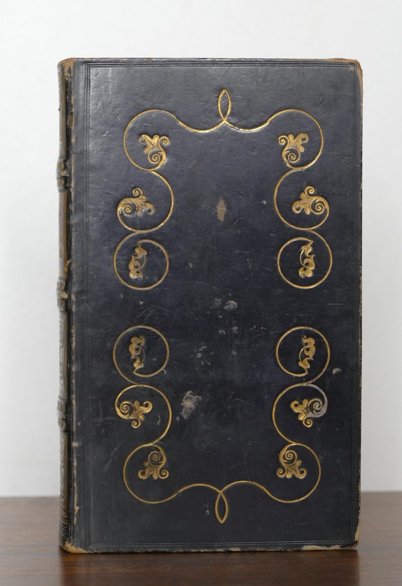 May include: Antique book with a dark blue leather cover. The cover features gold-colored ornate scrollwork and a decorative border. The spine is a lighter brown color. The book appears to be well-preserved and of historical significance.