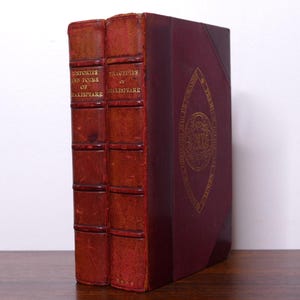 May include: Two antique books with leather covers. The spines are a reddish-brown color, with gold lettering that reads "Histories and Poems of Shakespeare" and "Tragedies of Shakespeare." The side of the book is a deep burgundy with gold circular design.