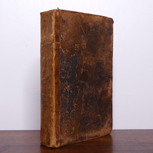 Puede incluir: Un libro antiguo, alto y rectangular, con una cubierta de cuero marrón desgastado. La cubierta muestra manchas oscuras y signos de edad, con un lomo visible y bordes ligeramente curvados. El libro está de pie sobre una superficie de madera.