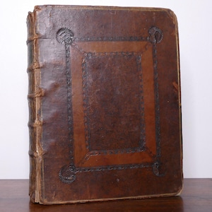 May include: A large, antique leather-bound book. The cover is a rich brown with a darker brown border and decorative corner accents. The spine shows the binding with raised bands. The book appears aged and well-preserved.