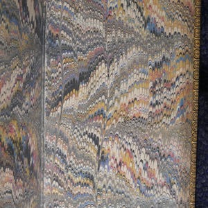 May include: Open book pages with marbled paper design. The paper features a swirling pattern in shades of gray, black, yellow, blue, and red. The edges of the pages are trimmed with a gold border.
