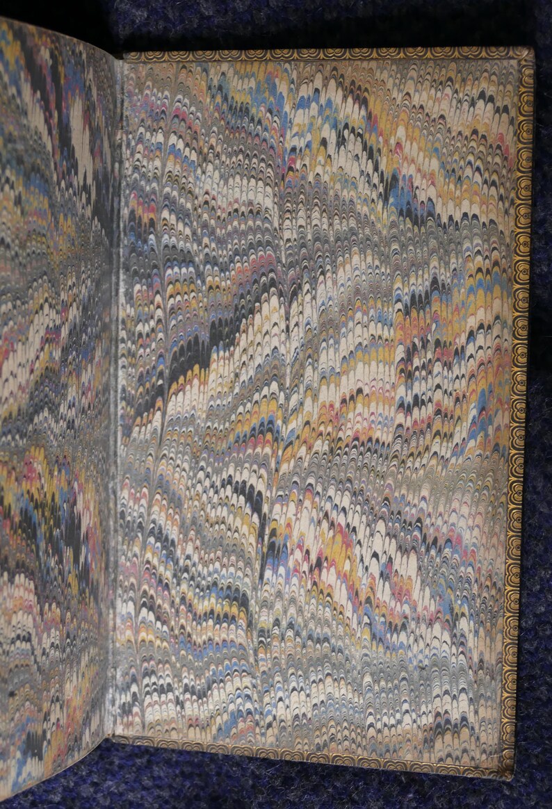 May include: Open book pages with marbled paper design. The paper features a swirling pattern in shades of gray, black, yellow, blue, and red. The edges of the pages are trimmed with a gold border.
