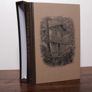 Pode incluir: Um livro encadernado em marrom com o título "Dickens na Europa" impresso em letras douradas na lombada. A capa apresenta uma ilustração em preto e branco de um edifício com uma varanda e árvores.