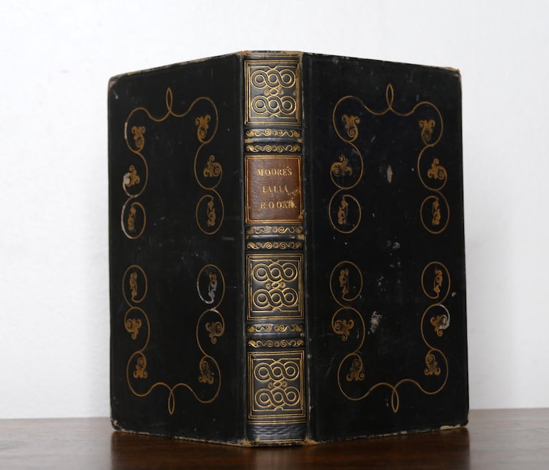 May include: A vintage book with a black cover and gold decorative accents. The spine features the title "MOORE'S LALLA ROOKH" in gold lettering. The cover has gold scrollwork and circular designs. The book is displayed on a wooden surface.