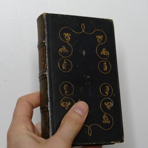 May include: A vintage, hardcover book with a black cover and gold decorative accents. The spine is brown with gold detailing. The book is held in a hand against a white background. The cover features ornate gold swirls and circular designs.
