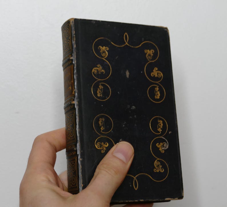 May include: A vintage, hardcover book with a black cover and gold decorative accents. The spine is brown with gold detailing. The book is held in a hand against a white background. The cover features ornate gold swirls and circular designs.