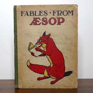 May include: A hardcover book titled Fables From Aesop with a tan cover featuring a red fox wearing glasses and reading a book. The title is printed in black and red lettering at the top, and the book has a green spine.