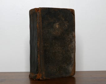 May include: A vintage, antique book with a dark brown leather cover. The book's edges show a contrasting orange-brown color, and the cover has a worn, textured appearance. The book is standing upright on a wooden surface.