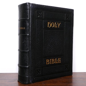 May include: A black leather-bound Holy Bible with gold lettering. The book is standing upright on a wooden surface. The spine has the word "BIBLE" in gold. The cover reads "HOLY BIBLE" in gold.