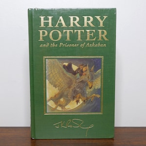 Puede incluir: Un libro de tapa dura verde titulado "HARRY POTTER and the Prisoner of Azkaban" con letras doradas. La portada presenta una ilustración de un grifo que lleva a una persona. La firma del autor está en la parte inferior.
