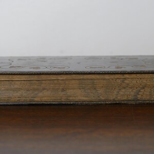 May include: A vintage book with a dark cover and aged, yellowed pages. The cover features embossed floral or circular designs. The book is closed and resting on a wooden surface, showcasing its age and historical charm.