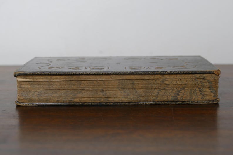 May include: A vintage book with a dark cover and aged, yellowed pages. The cover features embossed floral or circular designs. The book is closed and resting on a wooden surface, showcasing its age and historical charm.