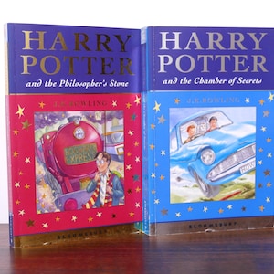 Puede incluir: Dos libros de la serie Harry Potter. "Harry Potter y la piedra filosofal" tiene una portada roja con una ilustración de un tren. "Harry Potter y la cámara secreta" tiene una portada azul con un coche volador.
