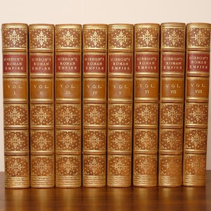 May include: A set of eight matching leather-bound books titled Gibbon's Roman Empire, numbered Volume I through Volume VIII. Each spine features a red label with gold lettering and intricate gold floral tooling on a tan leather background.